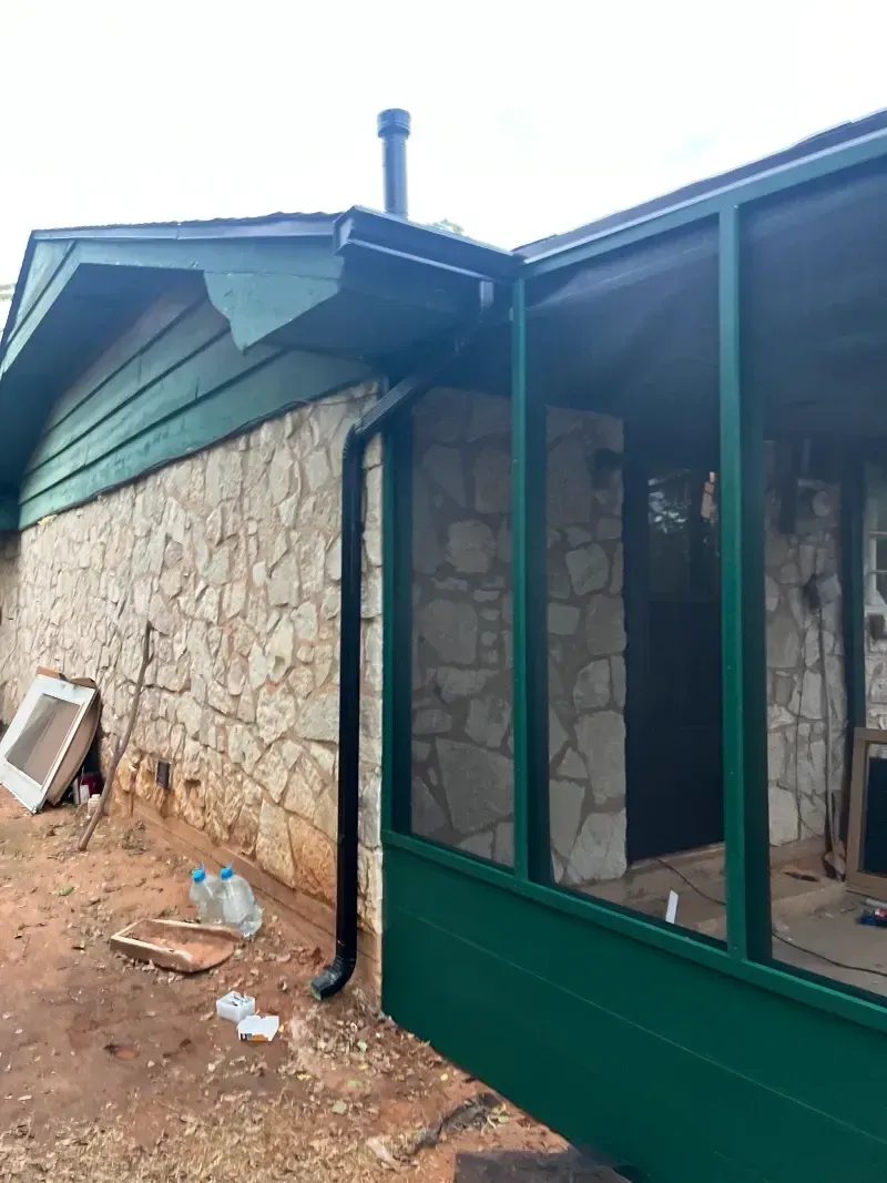 Exterior of a house with stone walls and a green screened porch. A black gutter and chimney are visible.