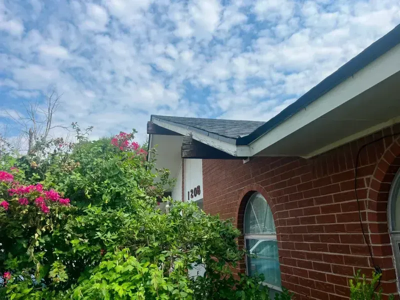 Red brick house with a dark roof and lush green bushes, under a cloudy sky.