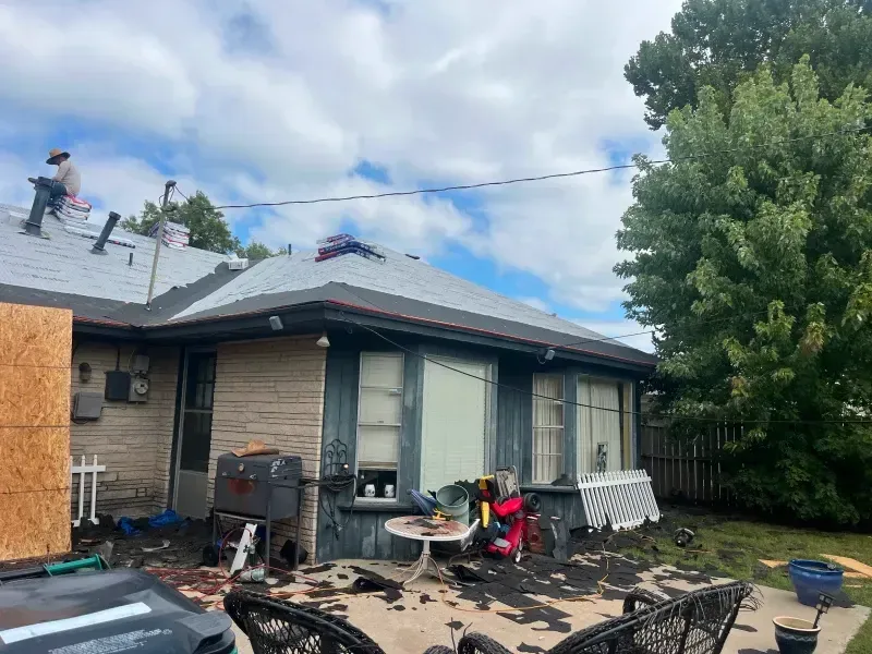 Person on a roof replacing shingles, debris scattered below. Brick house, blue-trimmed windows, cloudy sky.