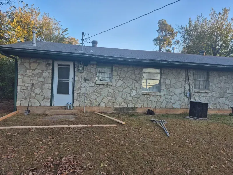 Stone-walled house with a door, windows, and a low, grassy yard. Boards and tools are in front of the house.