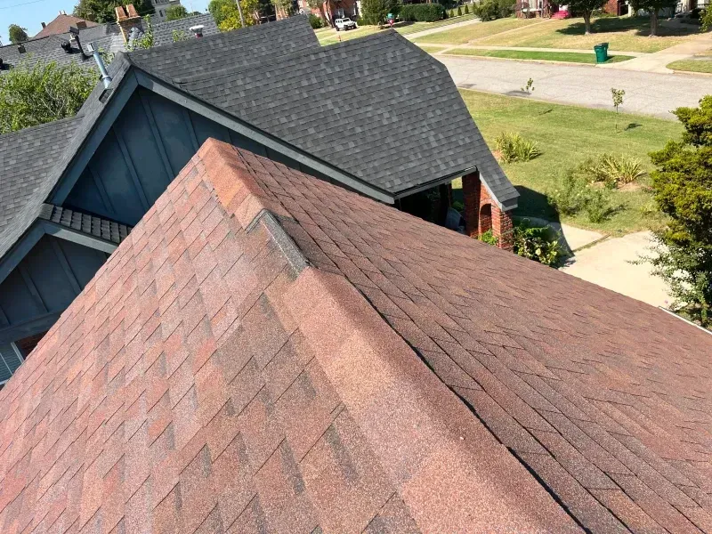 Two roofs with asphalt shingles, one red-brown, the other gray, on houses in a suburban setting.