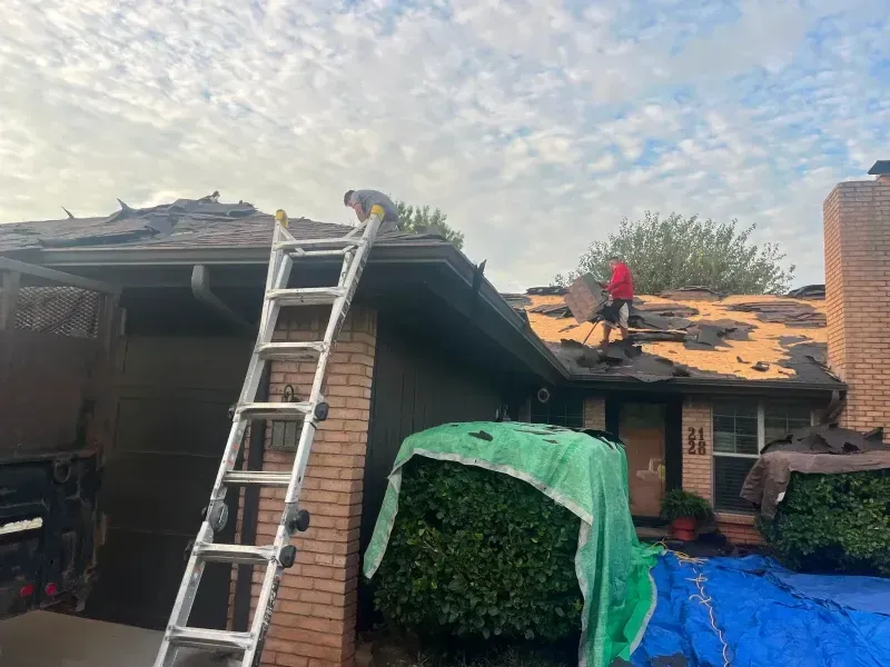 Workers replacing a damaged roof on a house, using a ladder. The sky is cloudy.