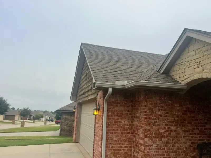 Brick house with brown shingle roof, tan garage door, and overcast sky.