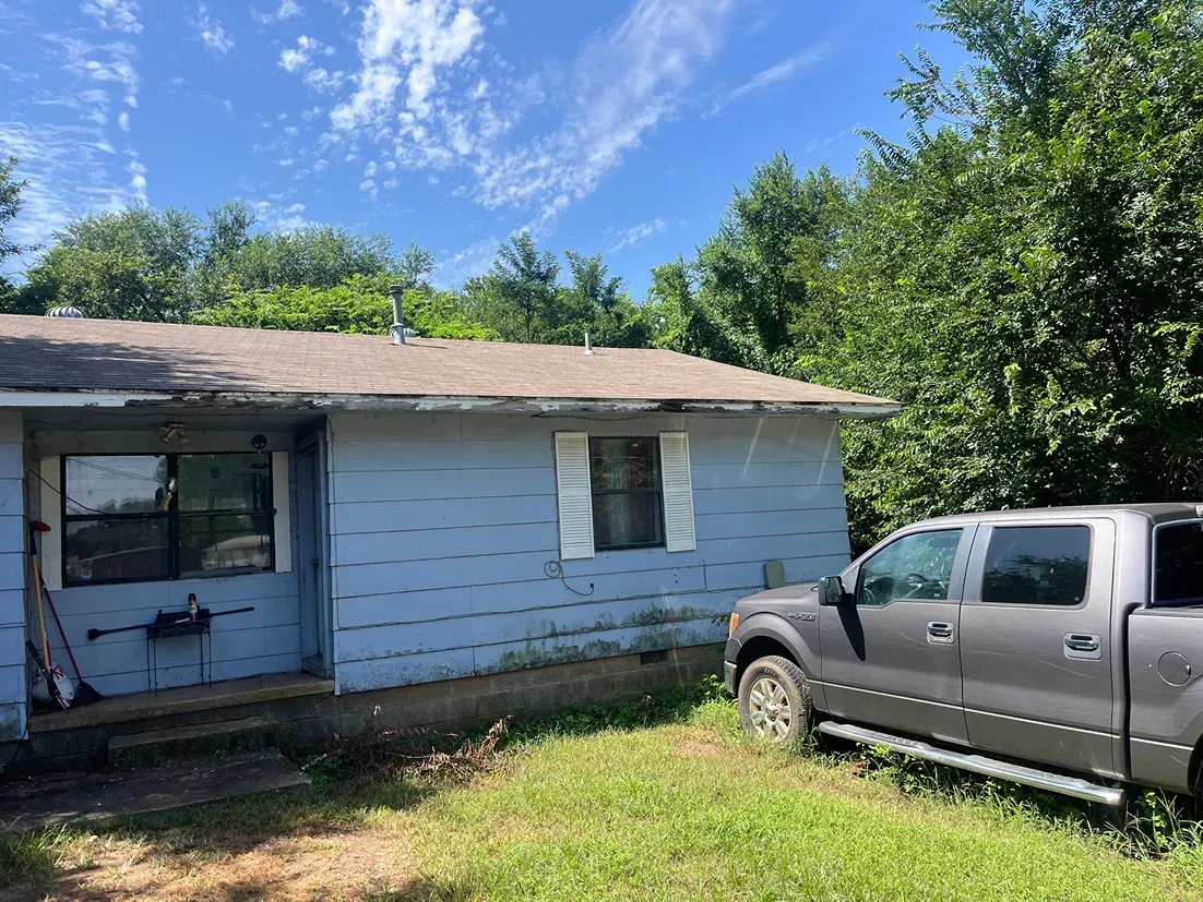 Blue house with a pickup truck parked beside it, surrounded by overgrown grass and trees.