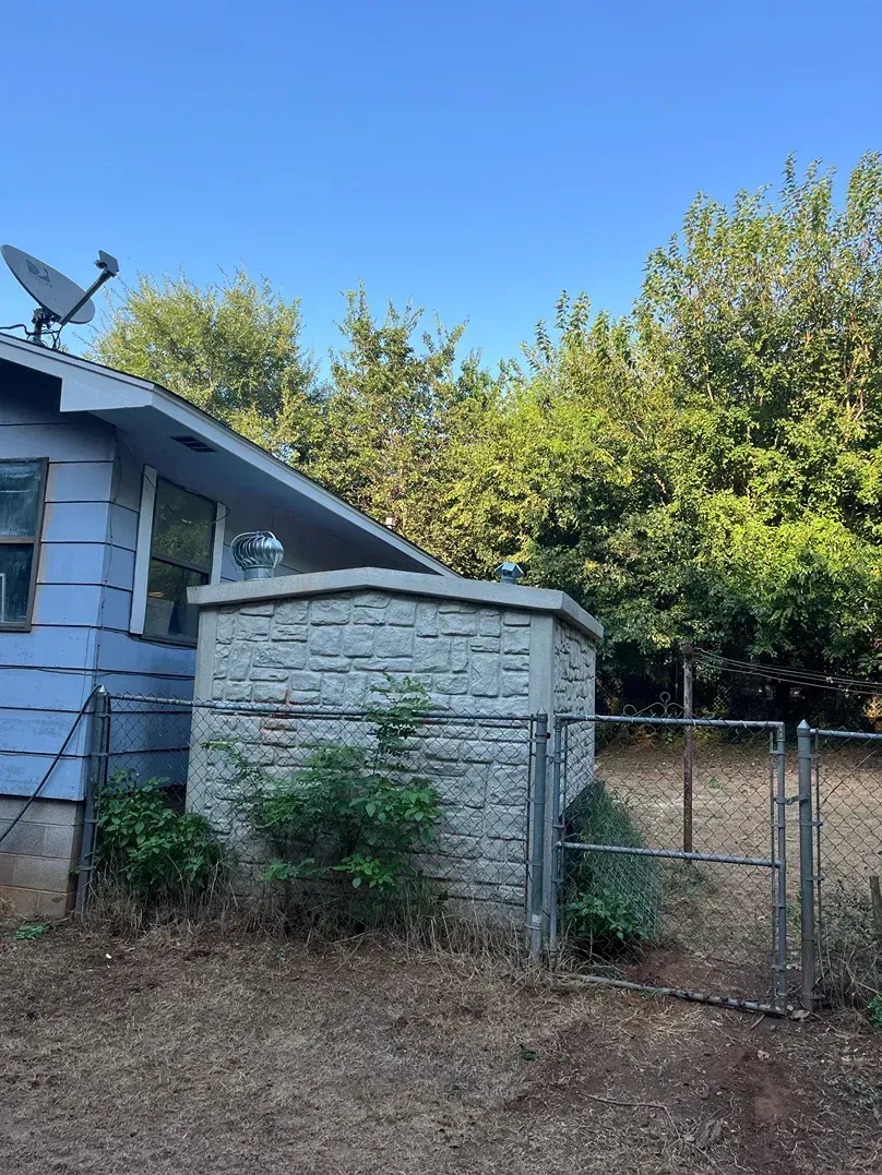 Small stone structure next to a light blue house, surrounded by trees and a chain-linked fence.