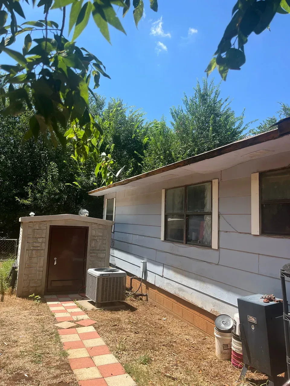 A light blue house with a brick path, shed, and an air conditioning unit. Trees in the background. Bright blue sky.