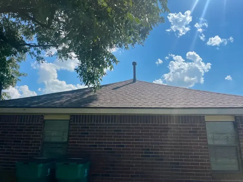 Brick building with brown roof, a metal vent pipe, and blue sky with clouds.