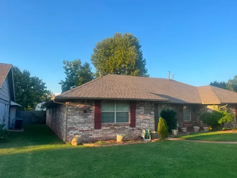 Brick house with brown roof, green lawn, trees, and blue sky.