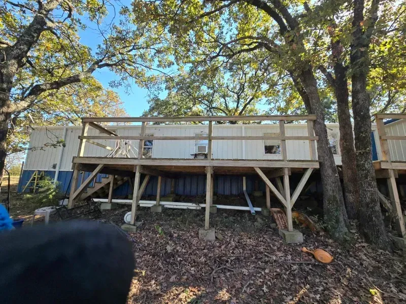 Wooden deck built onto a white building, under trees. Brown and blue siding below deck.