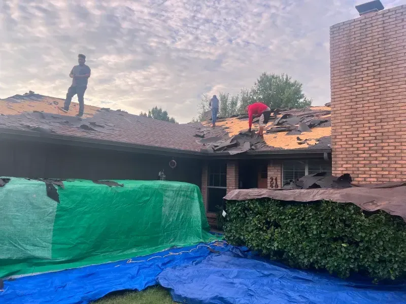 Roofing crew removing shingles from a house with a brick chimney; blue and green tarps cover portions of the house.