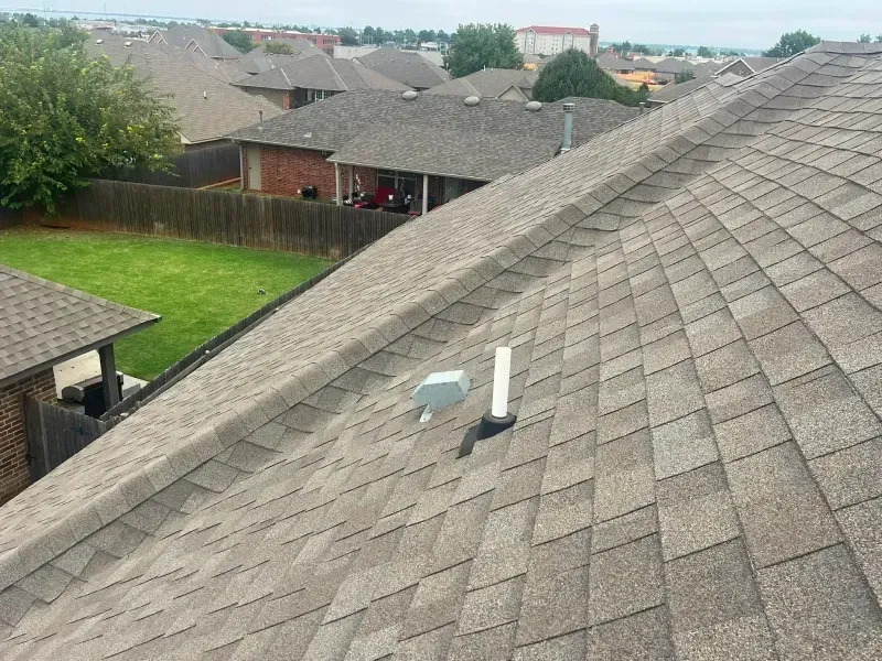 Brown shingled roof with a vent pipe on a suburban house. Houses and green lawn visible.