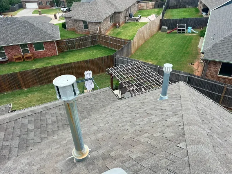 View of a brown shingled roof with three vent pipes. A backyard with green grass and fences is visible.