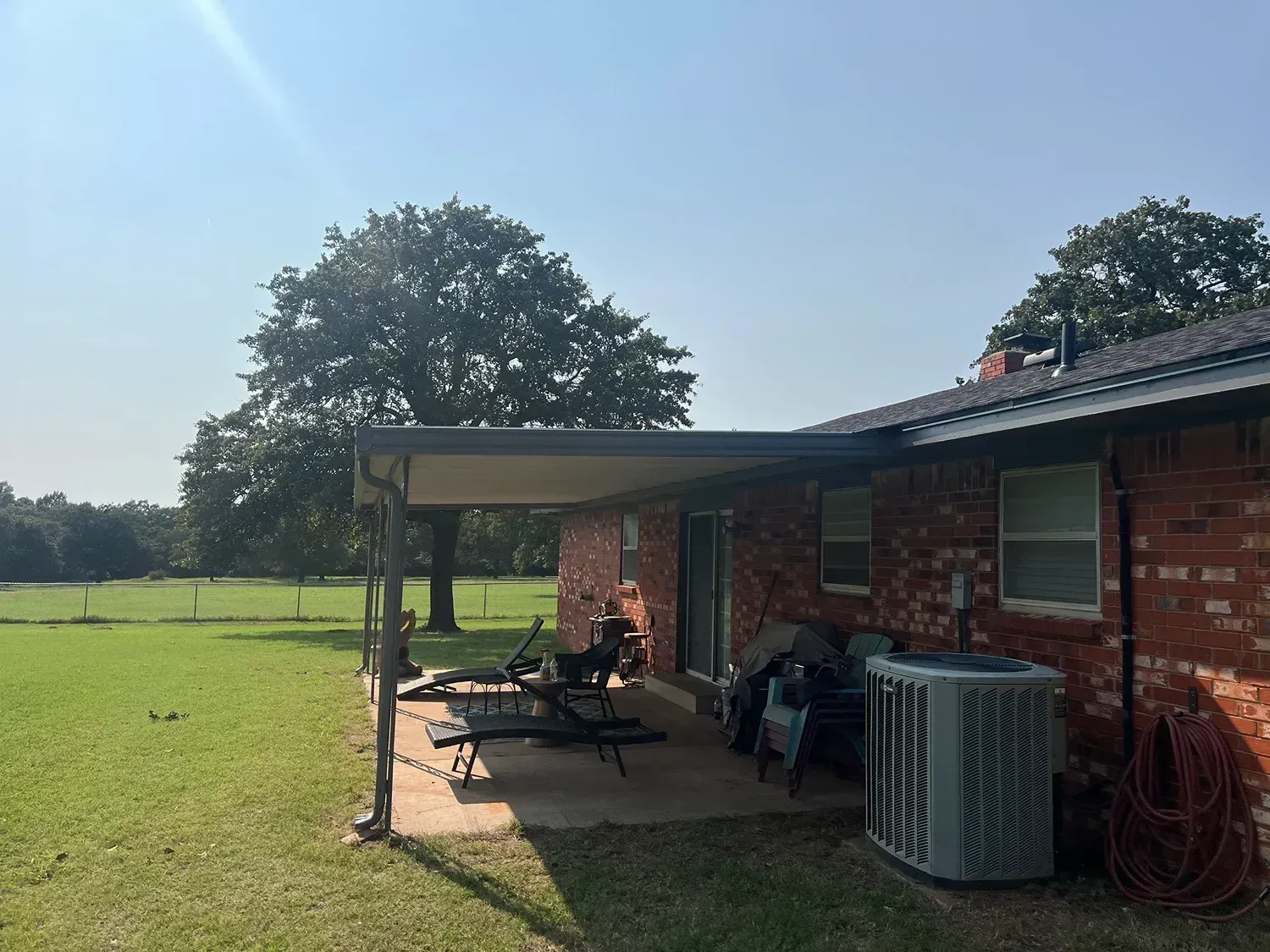 A red brick house with a patio roof, lawn, and large tree under a bright sky.