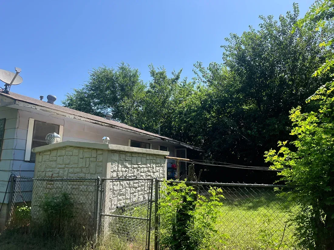 Side view of a house with a fence and overgrown vegetation.