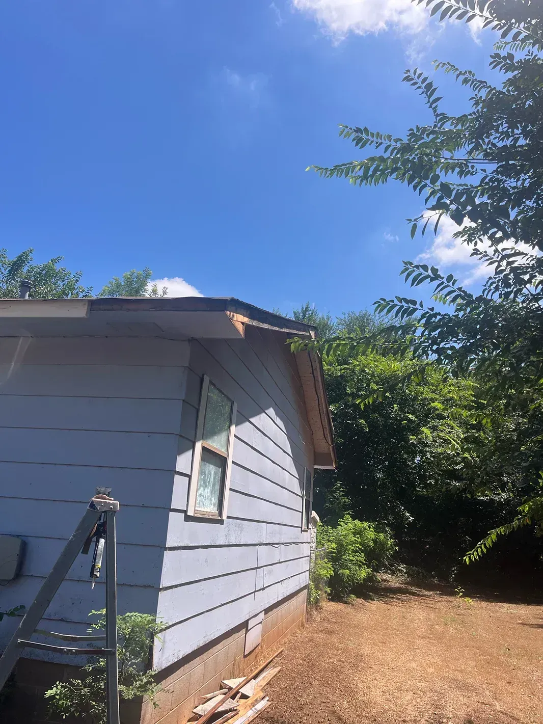Side view of a blue house with damaged roof and siding, next to trees, on a sunny day.