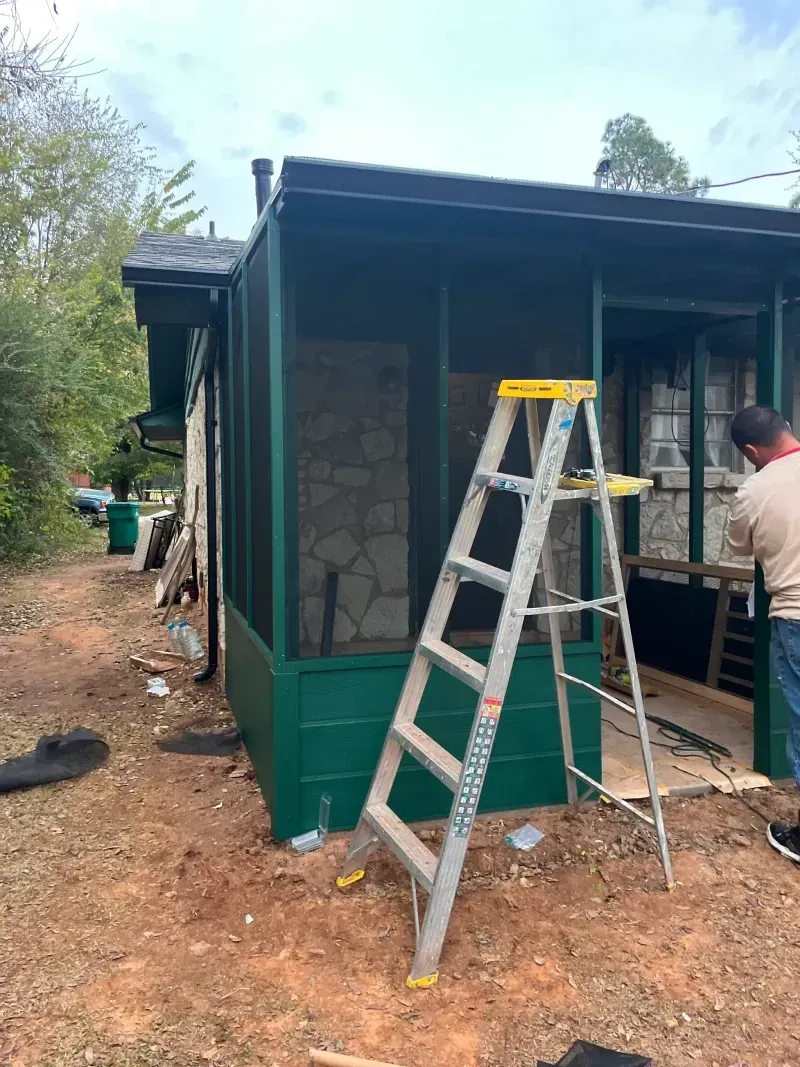A green screened-in porch under renovation, with a ladder and a person working on the window frame outside.