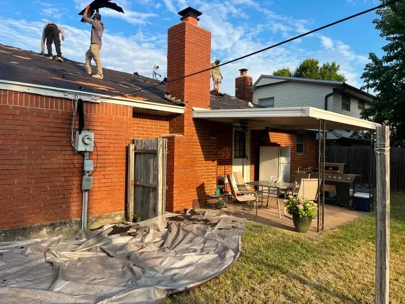 Roofers working on a brick house roof with a chimney; backyard with patio and tarp on ground.