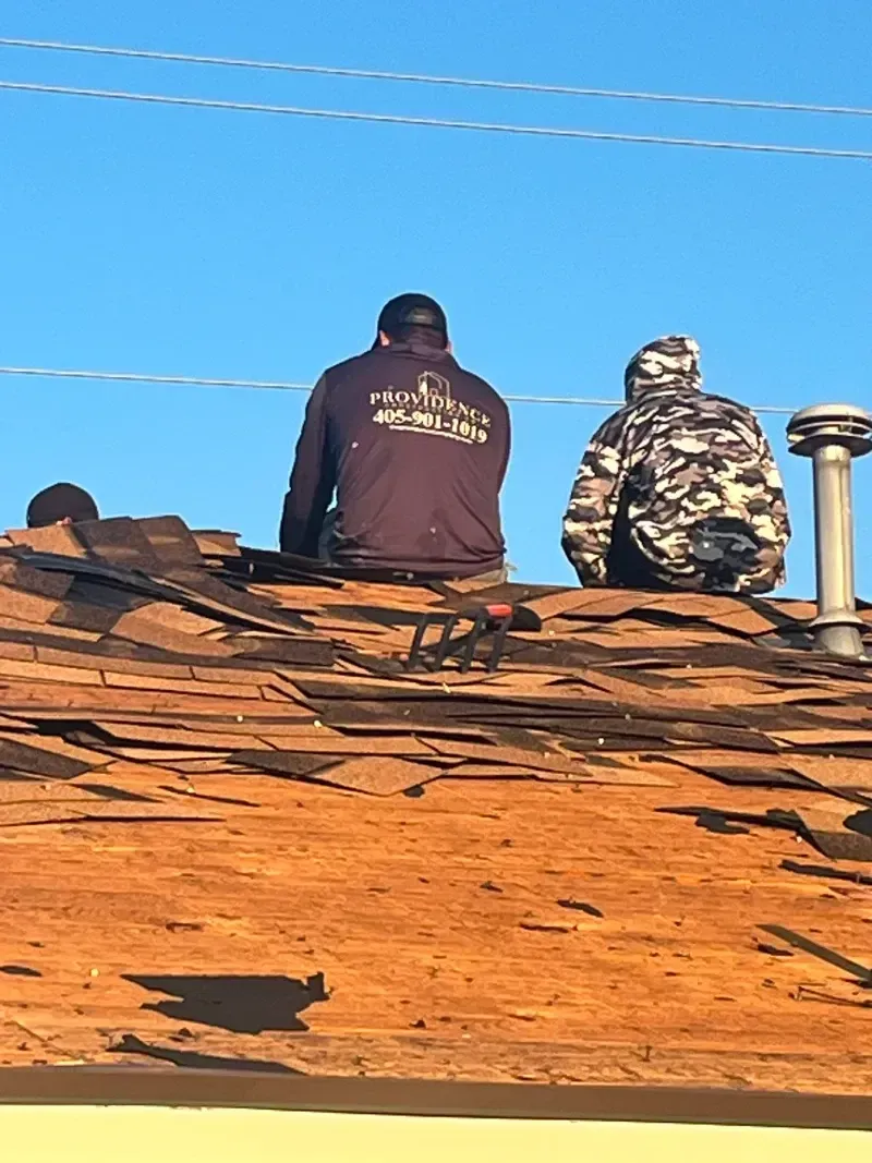 Two roofers sitting on a damaged roof under a clear blue sky, one wearing a jacket with company logo.