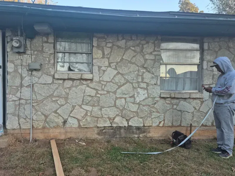 Person measuring the side of a stone-covered building. The windows have torn curtains.