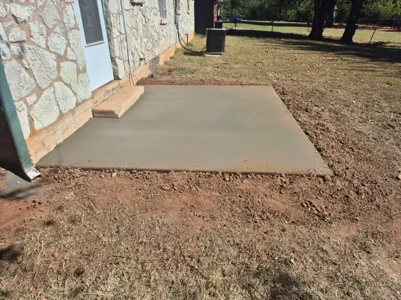 Newly poured concrete patio next to a stone building, with steps leading to a door. Brown dirt surrounds.