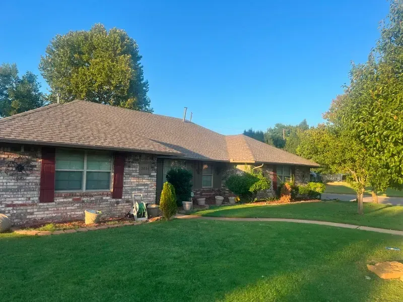 Single-story brick house with a brown shingled roof, green lawn, trees, and a clear blue sky.