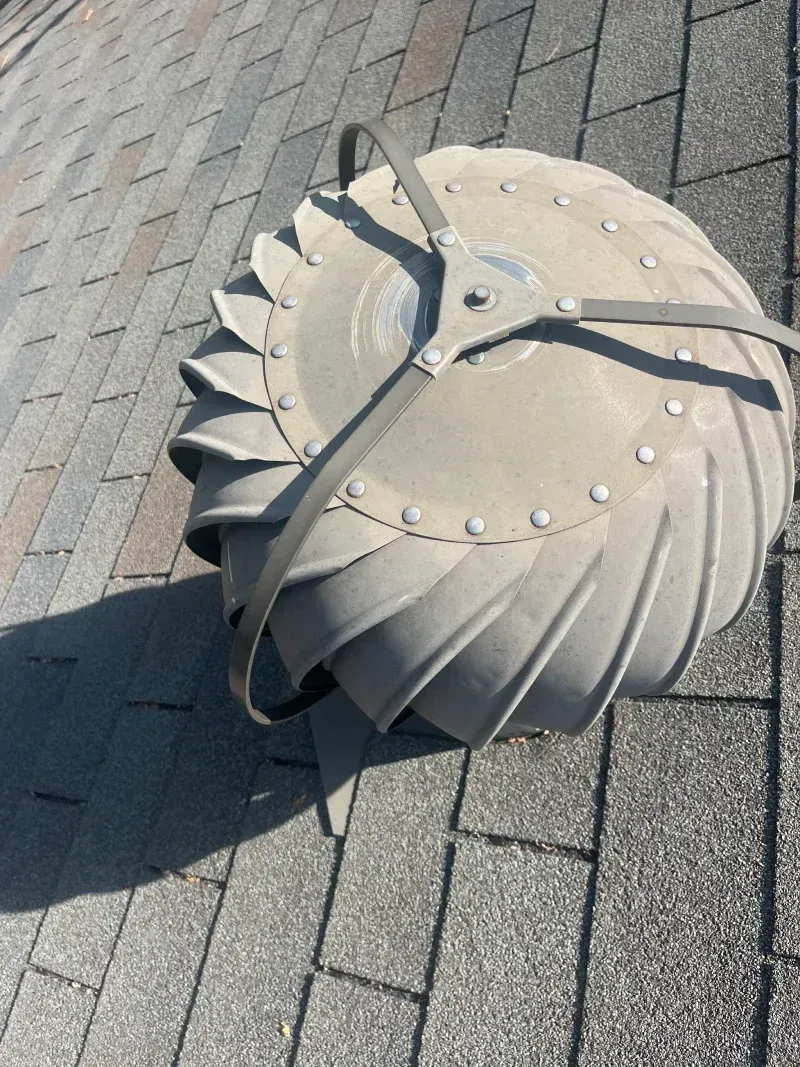 Rooftop turbine vent, metal, on asphalt shingles. Sunlight casts a shadow.