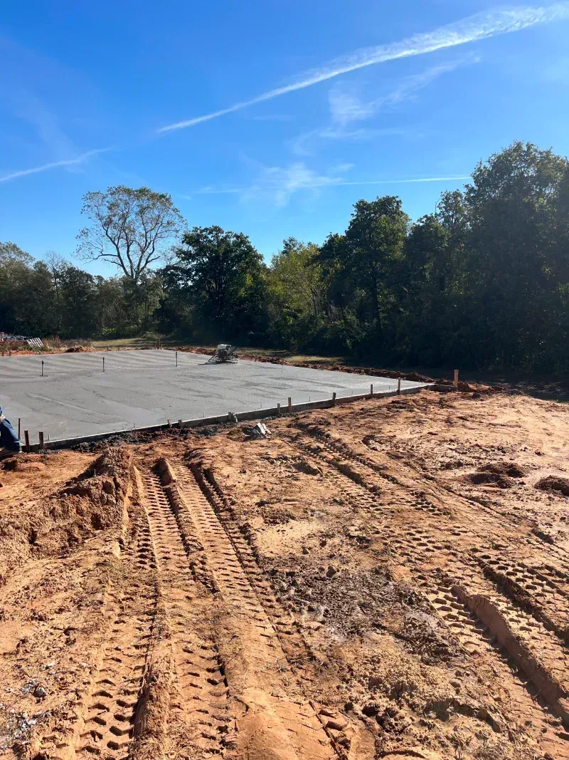 Concrete foundation on dirt ground with tire tracks under a blue sky, trees in the background.