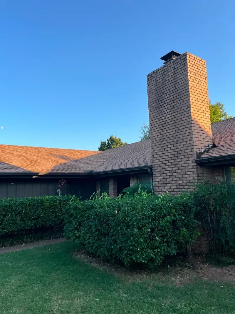 Brick chimney on a house with a tiled roof, green bushes, and lawn against a blue sky.