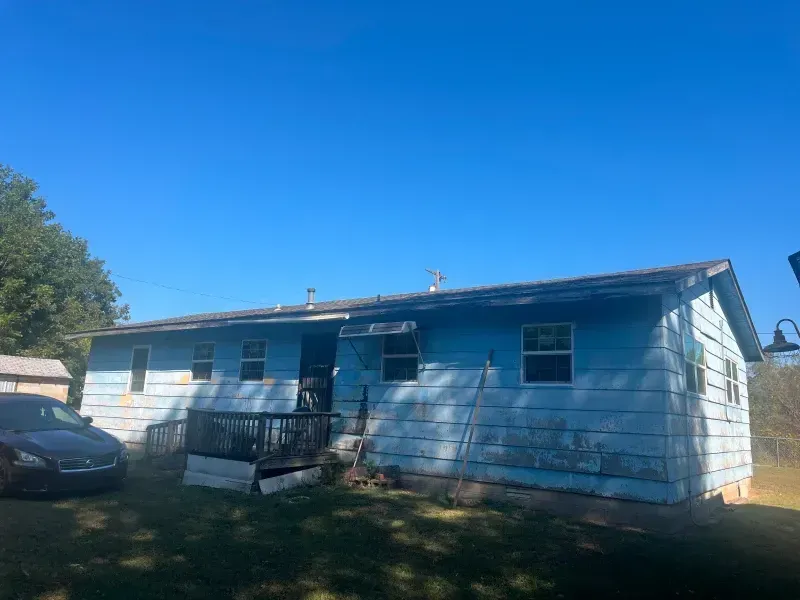 Blue-painted house with weathered siding. A black car sits nearby. Clear, blue sky.