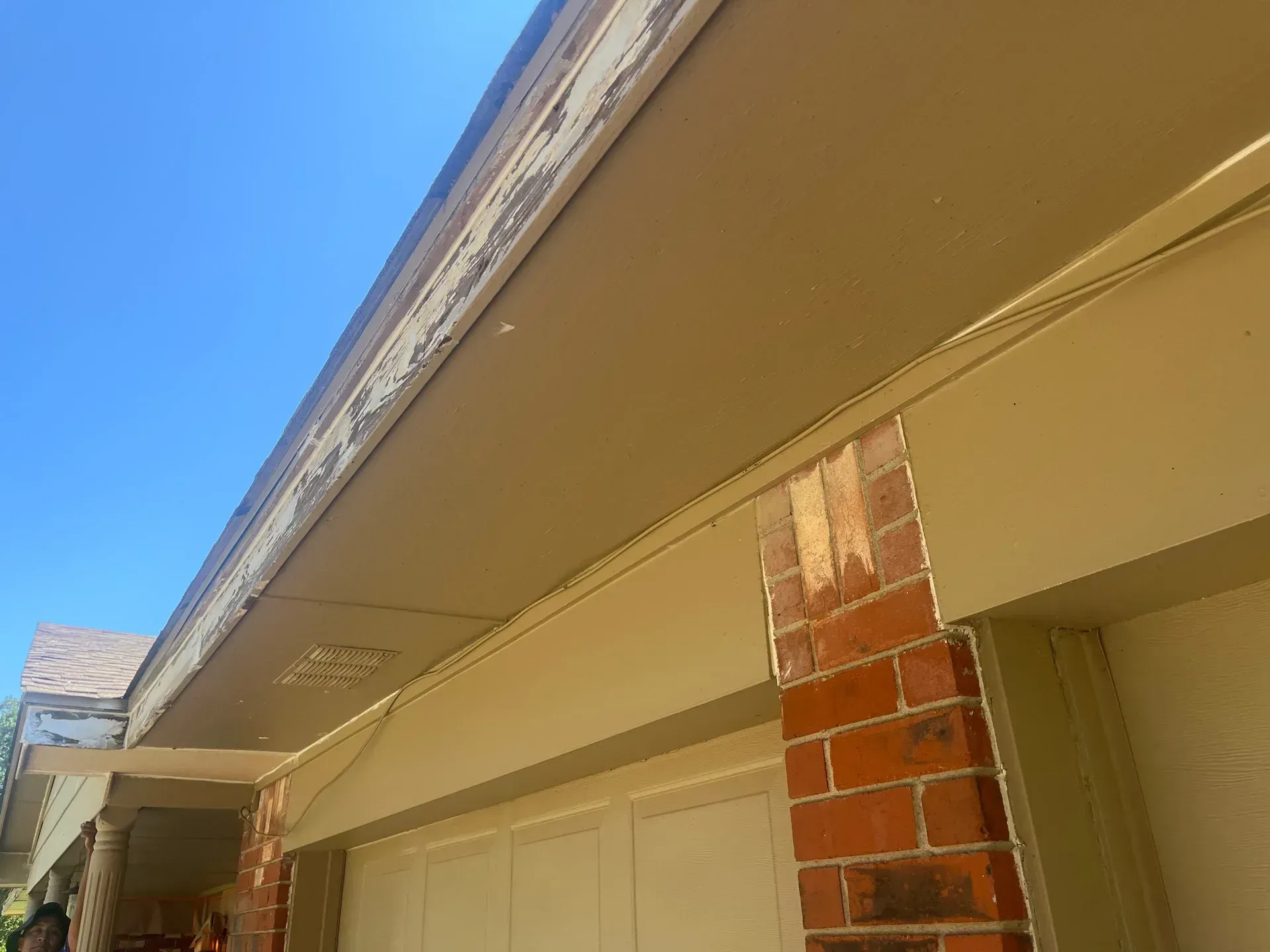 Exterior of a house with peeling paint on the eaves, brick and tan siding under a bright blue sky.