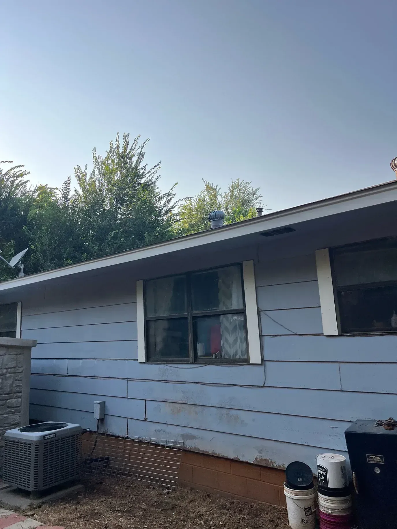 Blue house with white trim, windows, and an air conditioning unit. Trees in the background, clear sky.