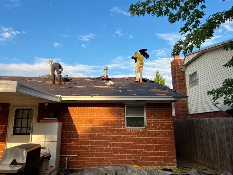Two people removing shingles from a brick house roof under a blue sky.