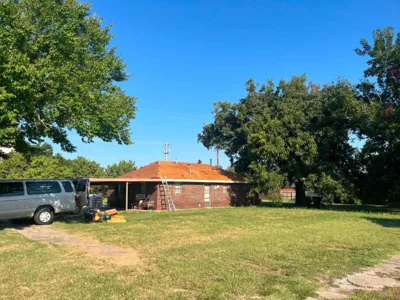 A one-story brick house with a brown roof and a grassy yard under a blue sky, a white van is parked on the left.