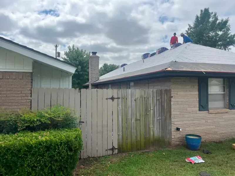 Two roofers on a house roof on a cloudy day, wooden fence in foreground.