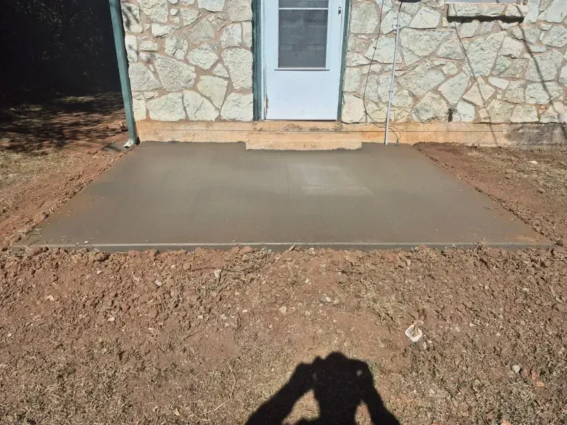 Concrete patio outside a stone-walled building. Brown earth surrounds the patio. Shadow of a person visible.