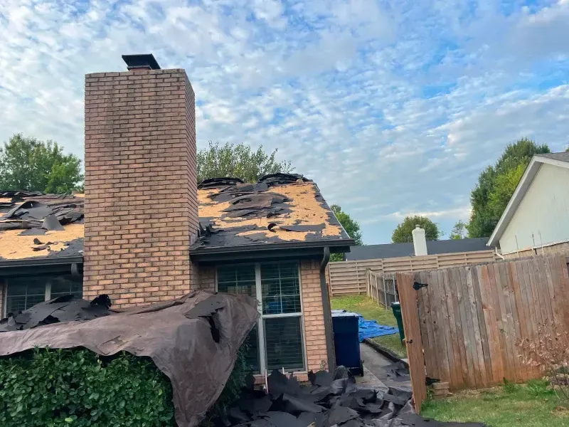 Damaged house roof with exposed underlayment and brick chimney, blue sky background.