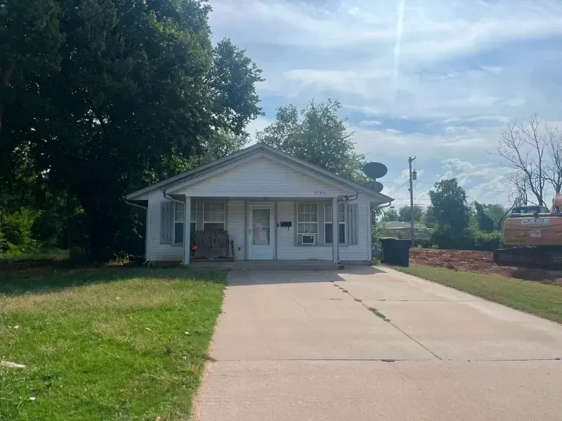 White house with a concrete driveway and grass lawn. Excavator and trees in the background. Blue sky.