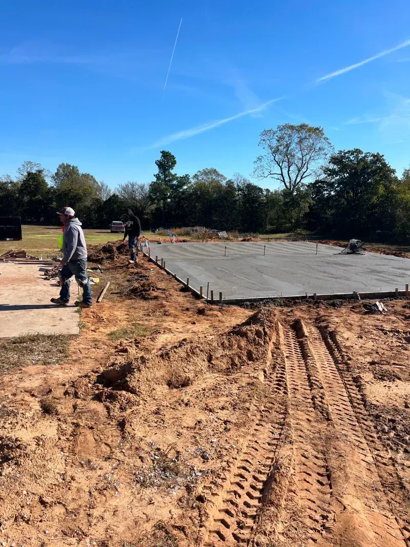 Construction site: workers near a concrete foundation on a sunny day. Dirt and tire tracks in foreground.