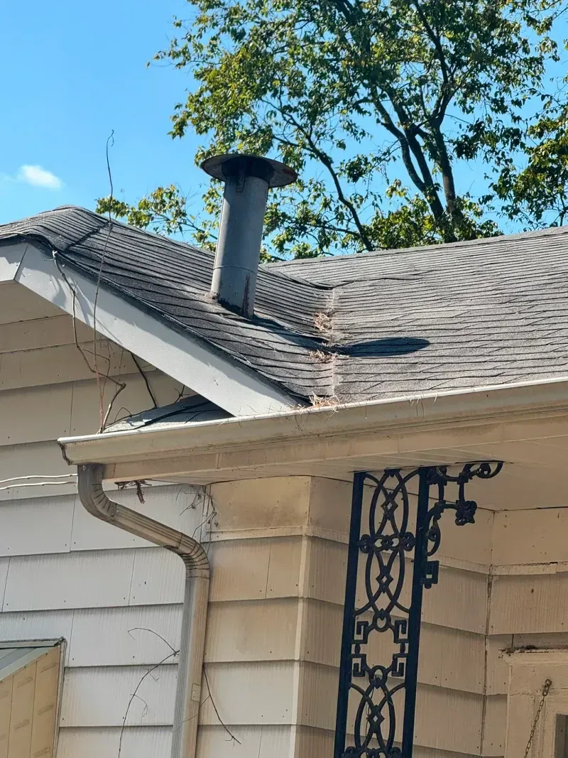 A house roof with a chimney, gutter, and decorative porch detail against a blue sky with tree foliage.