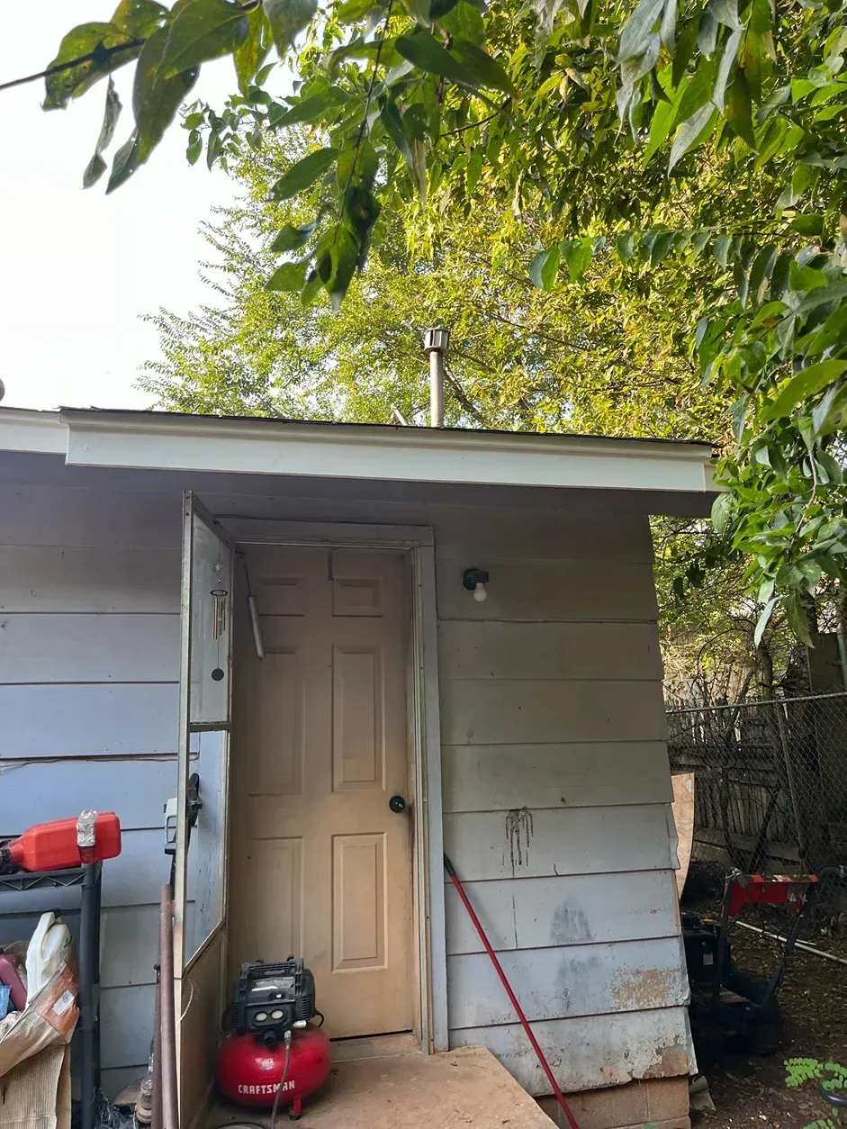 A small, light gray shed with a closed door, red air compressor, and a metal chimney.