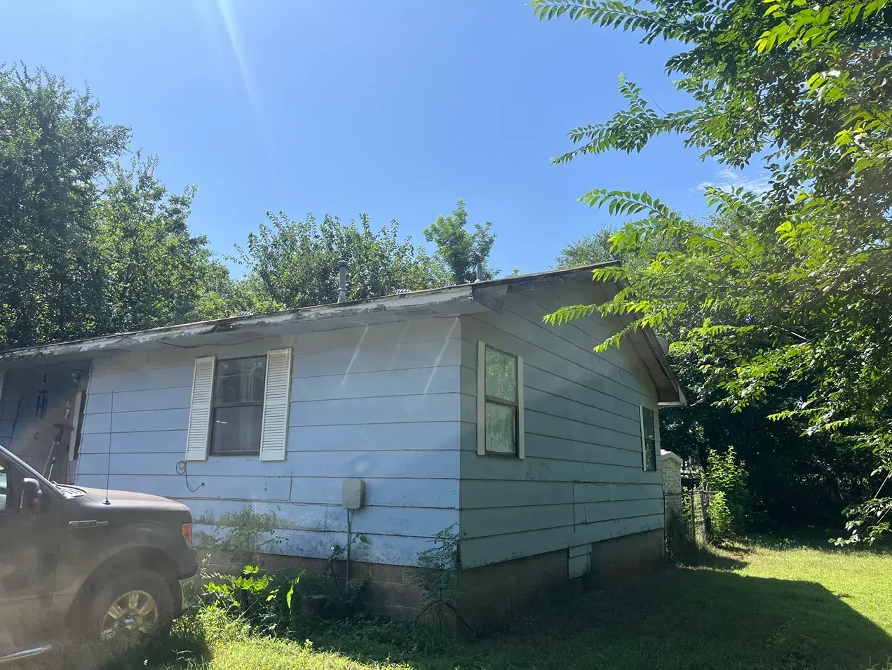 Side view of a blue-sided, single-story house with a metal roof, set on a grassy lot, partially obscured by trees.