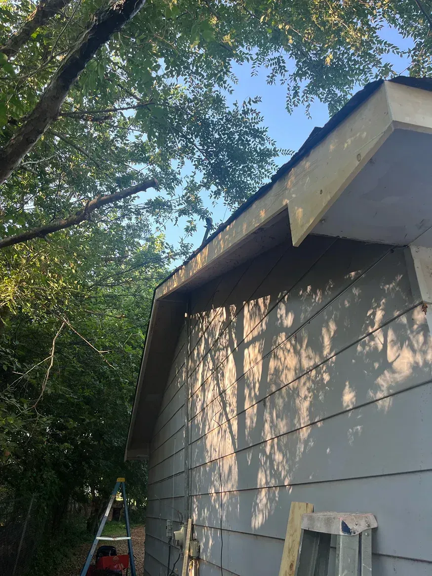 Side of a gray building with a newly constructed roof overhang, against a backdrop of trees and a blue sky.