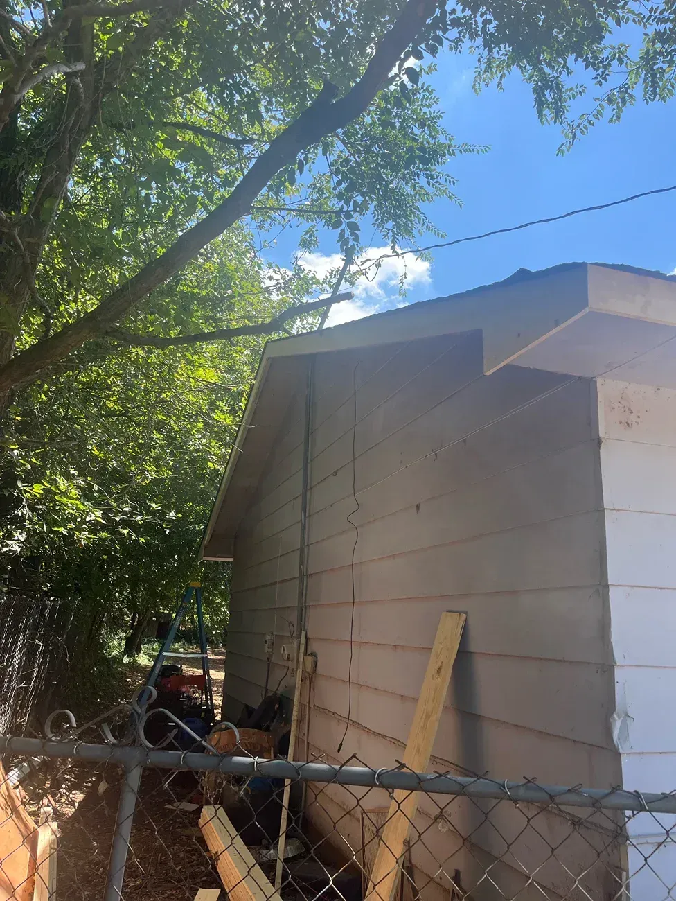 Side of a building with light-colored siding, under a blue sky, partially obscured by green trees.