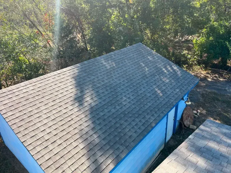 A gray shingle roof on a blue building, surrounded by green trees in sunlight.