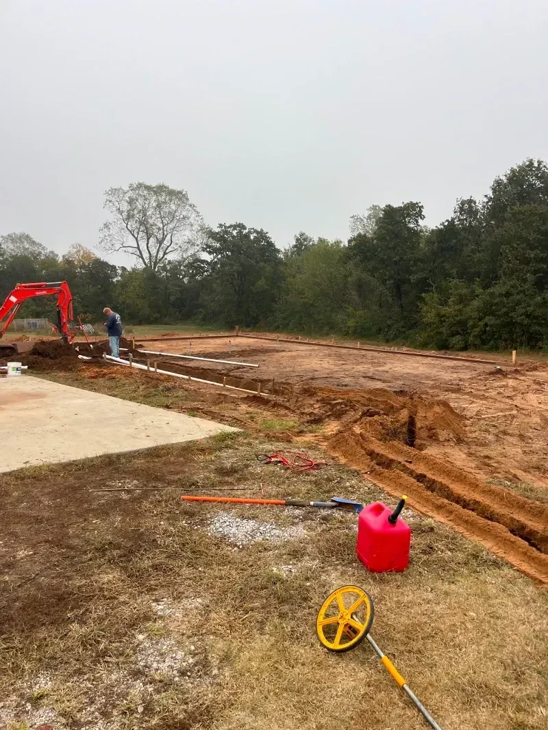 Construction site with a backhoe digging trenches in a dirt field. A person stands near a trench.