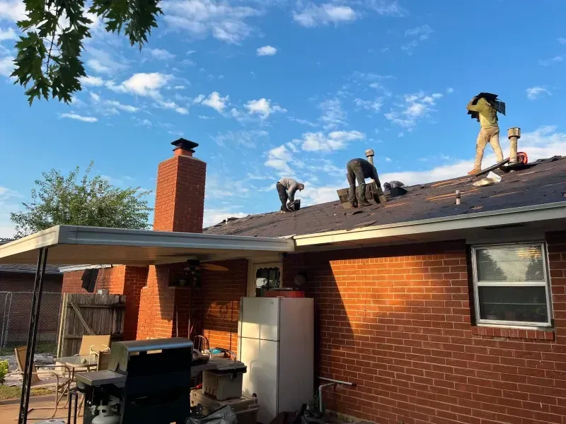 Roofers working on a residential roof under a partly cloudy blue sky. Brick house with a chimney and awning.