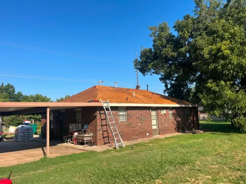 Brick house with reddish-brown roof, carport, and ladder; on a grassy yard under a blue sky.