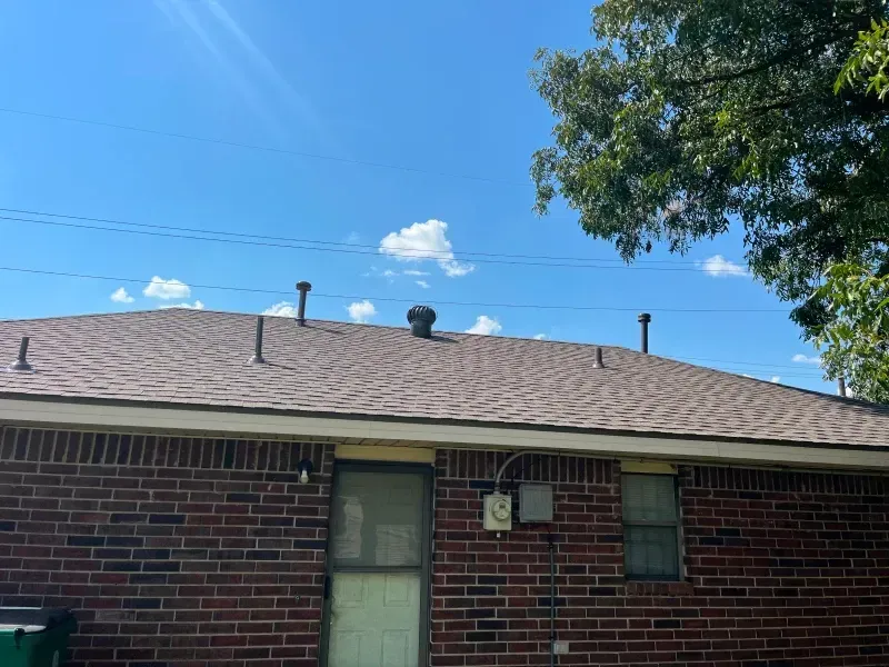 Brick building with a brown shingle roof, vents, and blue sky with clouds.