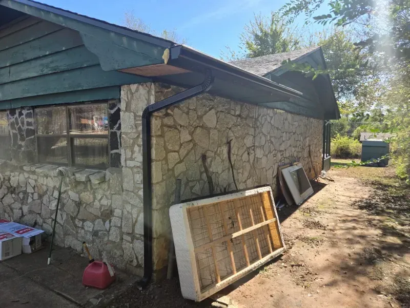 Stone-walled building with green trim and a black gutter. A mattress and other items are outside.