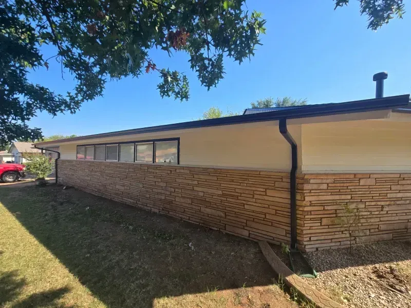 A one-story house with stone veneer, cream siding, black gutters, and windows under a blue sky.